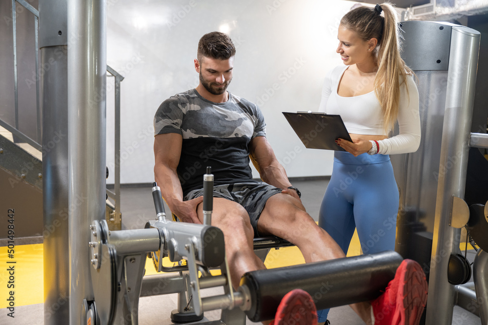 Obraz premium Muscular handsome man doing exercises on leg press machine and female personal trainer with clipboard calculating his muscle progress during workout class at gym.