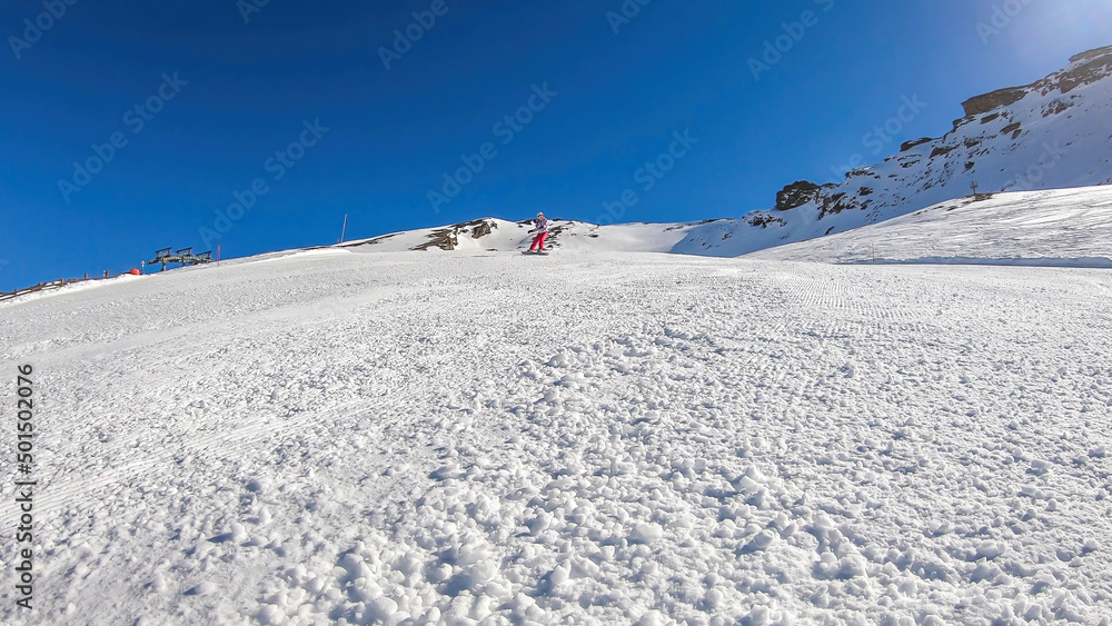 A snowboarder going down the slope in Heiligenblut, Austria. Perfectly groomed slopes. High mountains surrounding the man wearing yellow trousers and blue jacket. Girl wears helm for the protection.