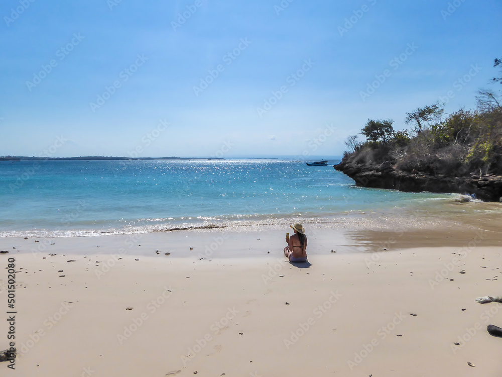 A girl in bikini and straw hat sitting on Pink Beach, Lombok Indonesia. Girl is enjoying the heavenly view. The water has many shades of blue. The beach is surrounded by small hills. Paradise beach.