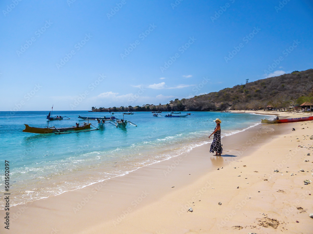 A woman in maxi dress walking on the Pink Beach in Lombook, Indonesia. Beautiful beach. Bay full of boats. No other people. Beautiful colors of the sand and the water. Dried trees in the background.