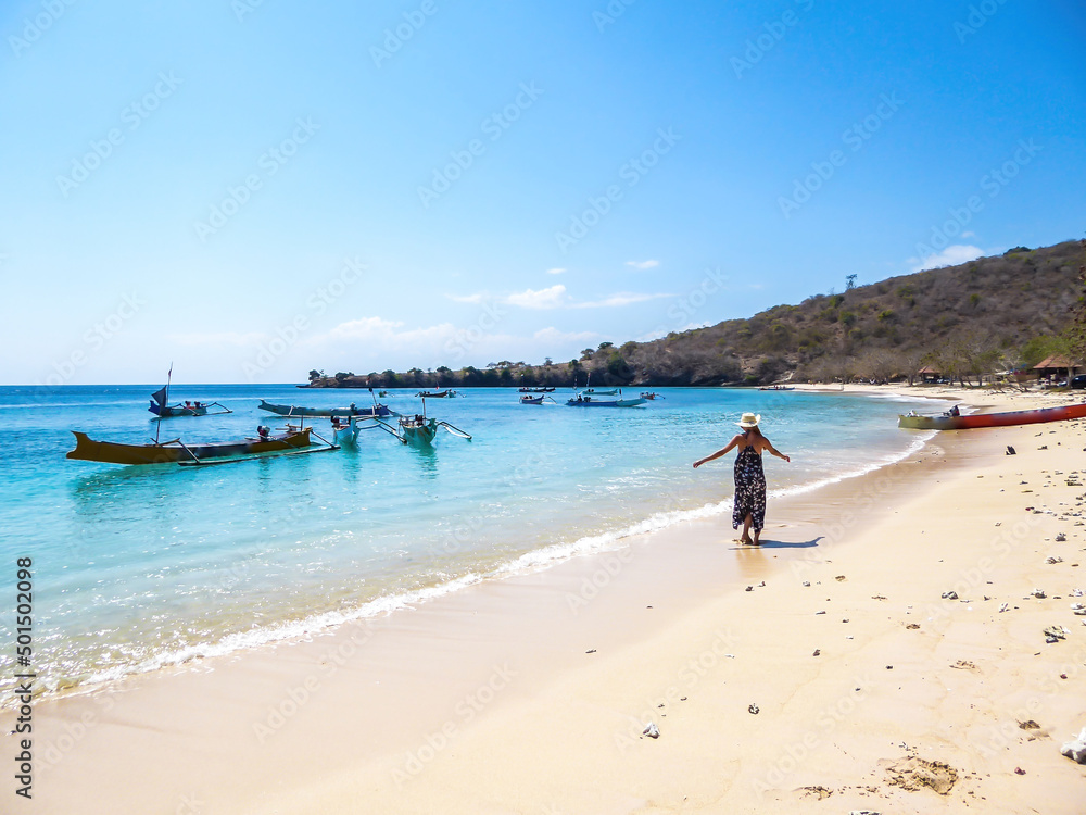 A woman in maxi dress walking on the Pink Beach in Lombook, Indonesia. Beautiful beach. Bay full of boats. No other people. Beautiful colors of the sand and the water. Dried trees in the background.