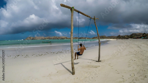 A man swinging on a swing placed on the seashore of Tanjung Aan Beach, Lombok, Indonesia. The swing has simple wood construction. Waves wash the pillars of it. In the back there are few boats.