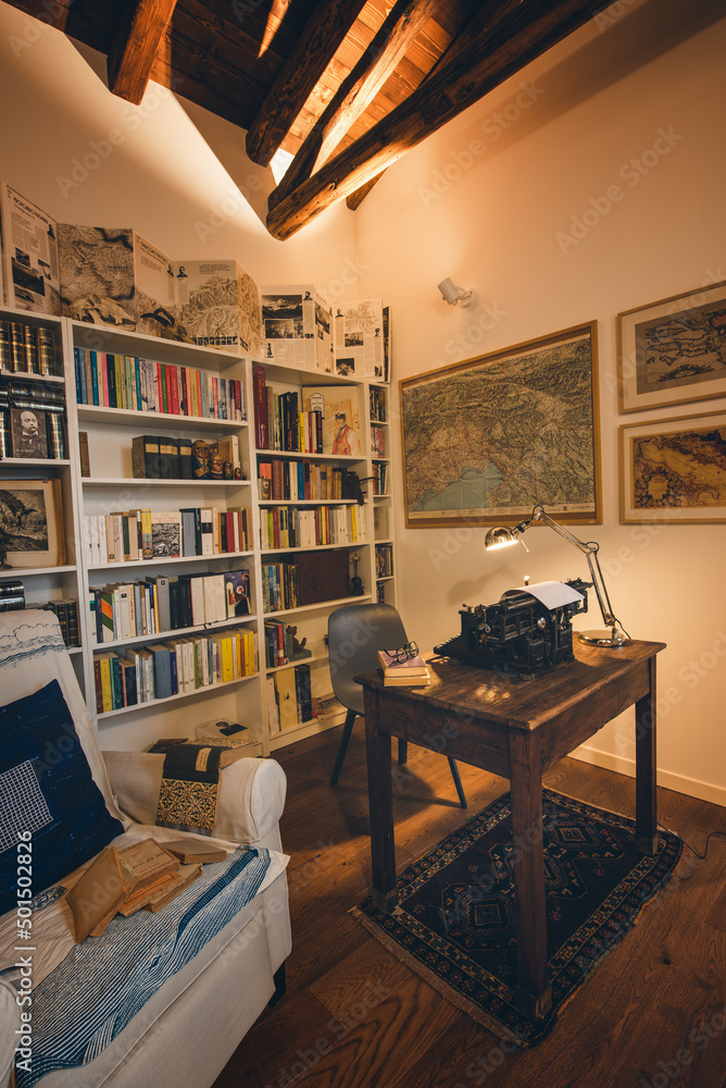 An old classic typewriter in a vintage style studio with a wooden desk lit by a lamp, wit old books all around. A moody, dark atmosphere for a writer working at night.