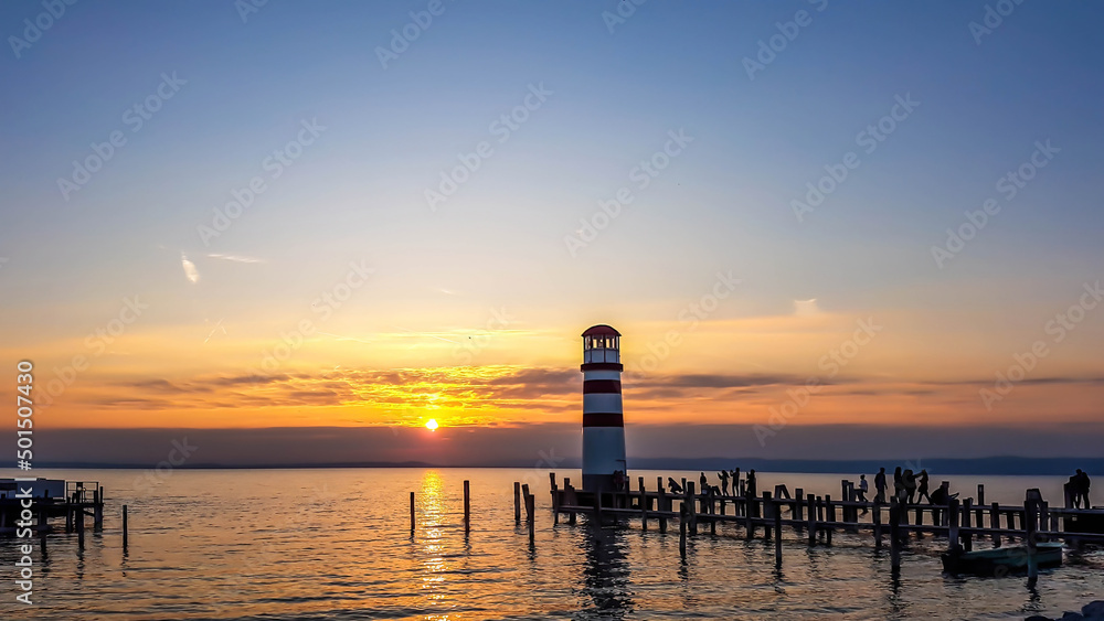 White and red striped lighthouse in Neusiedlersee, on Austrian-Hungarian border. Sun sets over the horizon in the clouds. Sky is orange, lake surface is soft and tender. Few clouds on the sky.