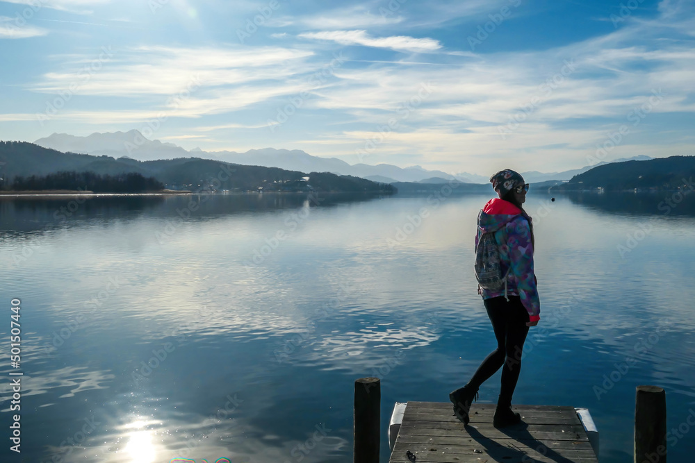 Girl wearing a jacket and beanie stands at the end of a promenade on the lake. Soft reflections of the clouds in the lake. Clear but cloudy day. High mountains in the back Calmness and relaxation