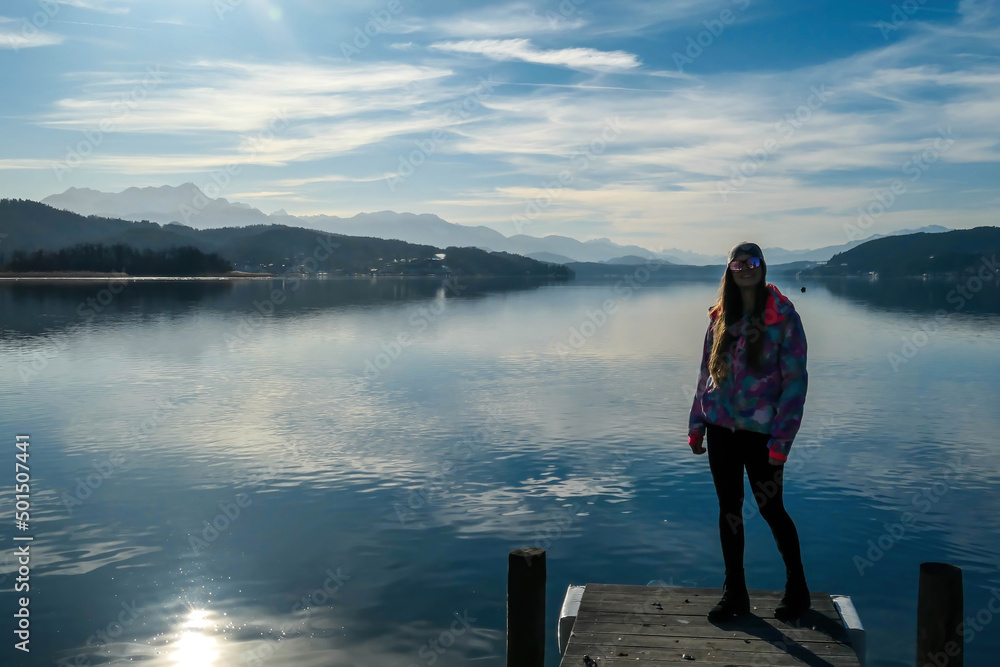 Girl wearing a jacket and beanie stands at the end of a promenade on the lake. Soft reflections of the clouds in the lake. Clear but cloudy day. High mountains in the back Calmness and relaxation