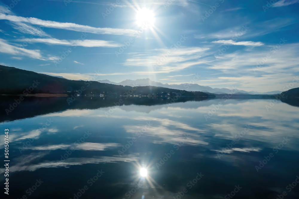A view on a lake and Alps in the back. The calm surface of the lake is reflecting the mountains, sunbeams and clouds. Clear and sunny day. Calm and relaxed feeling.