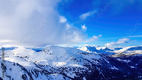 A winter landscape of mountains. Sun is shining up in the sky. Mountains slopes covered completely with thick layer of snow. Few trees on the slopes. The slopes look unfavorable and dangerous.