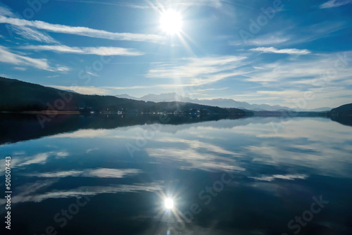 A view on a lake and Alps in the back. The calm surface of the lake is reflecting the mountains, sunbeams and clouds. Clear and sunny day. Calm and relaxed feeling.