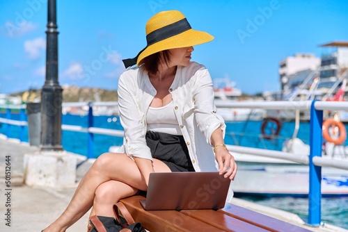 Outdoor, middle aged woman in straw hat sitting on bench using laptop