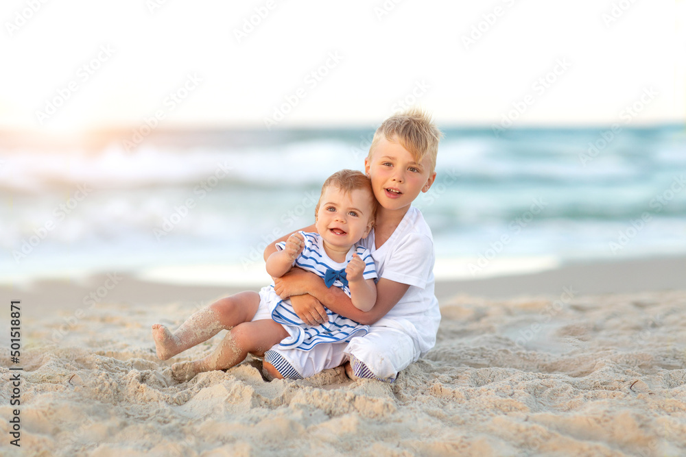 Smiling children sit on beach during summer holidays vacation. White and marine clothes. Toddler and kid, beautiful sea, sand and sunset.