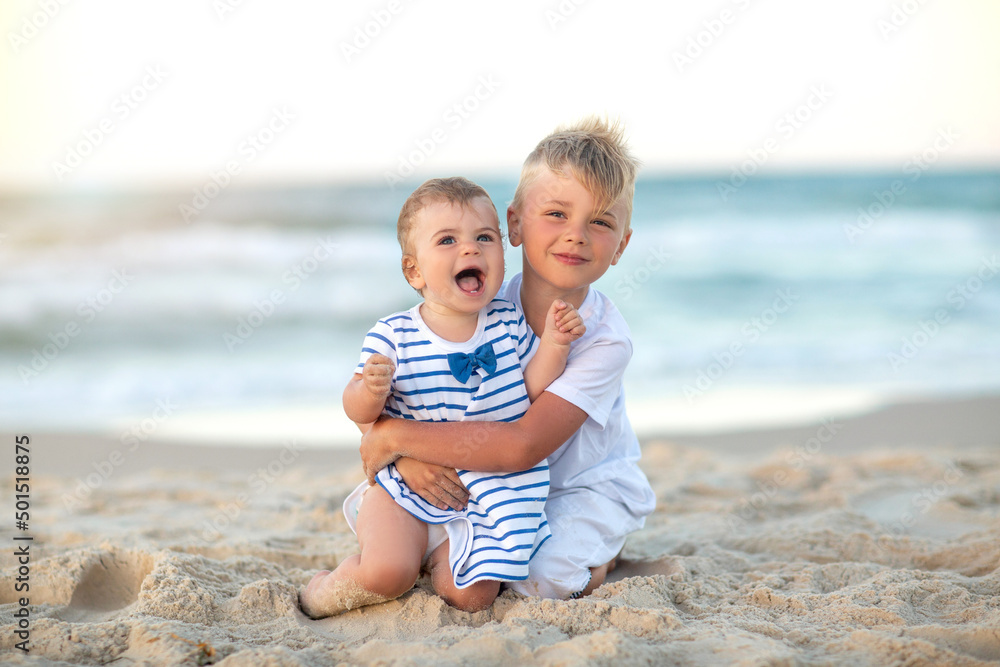 Smiling children sit on beach during summer holidays vacation. White and marine clothes. Toddler and kid, beautiful sea, sand and sunset. Smiling children sit on beach during summer holidays vacation. White and marine clothes. Toddler and kid, beautiful sea, sand and sunset.