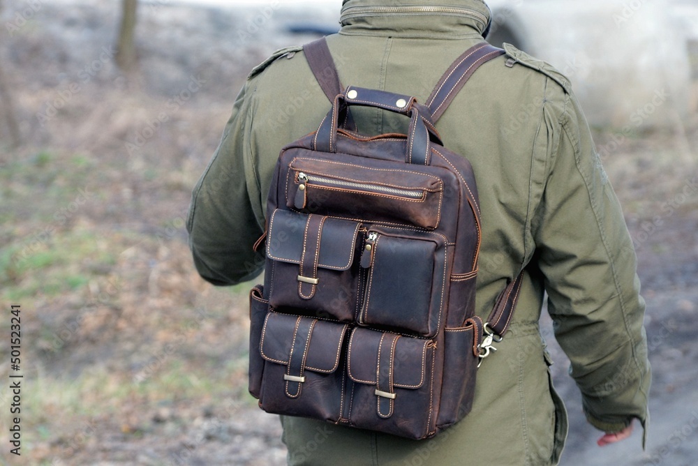one brown leather military backpack on the back of a man in a green jacket on the street
