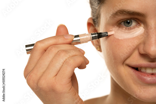 Half portrait of a young smiling woman applyes concealer under her eyes on white background