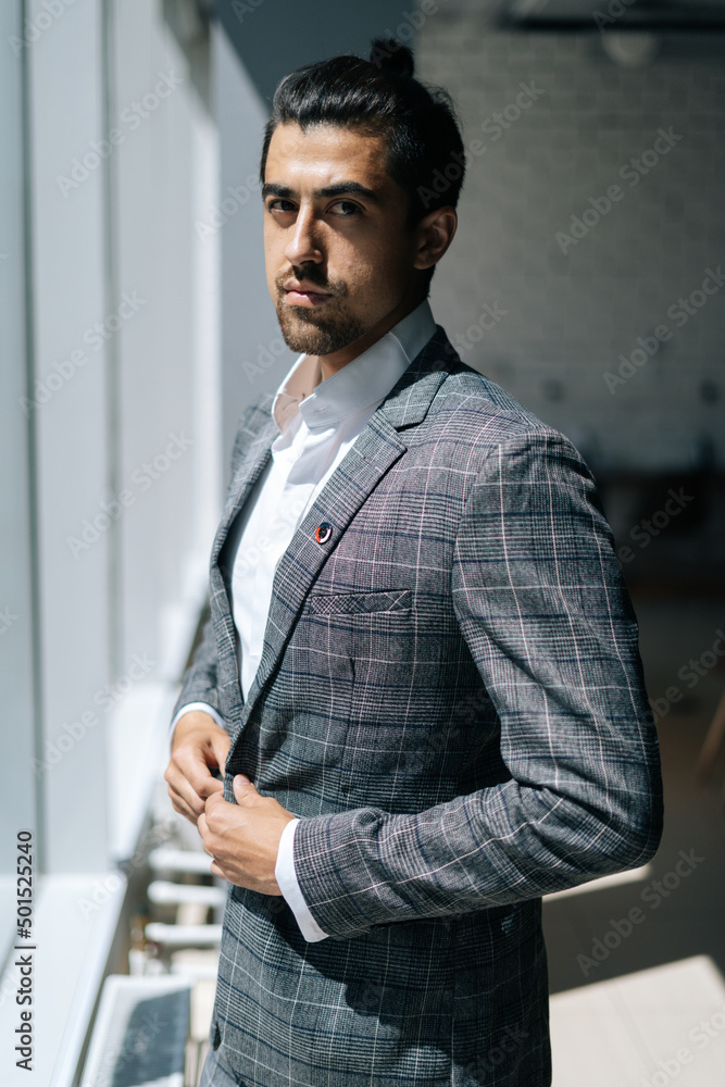 Vertical portrait of stylish handsome business man wearing fashion suit standing near window in modern office room, looking at camera. Front view of bearded businessman posing at workplace. Vertical portrait of stylish handsome business man wearing fashion suit standing near window in modern office room, looking at camera. Front view of bearded businessman posing at workplace.