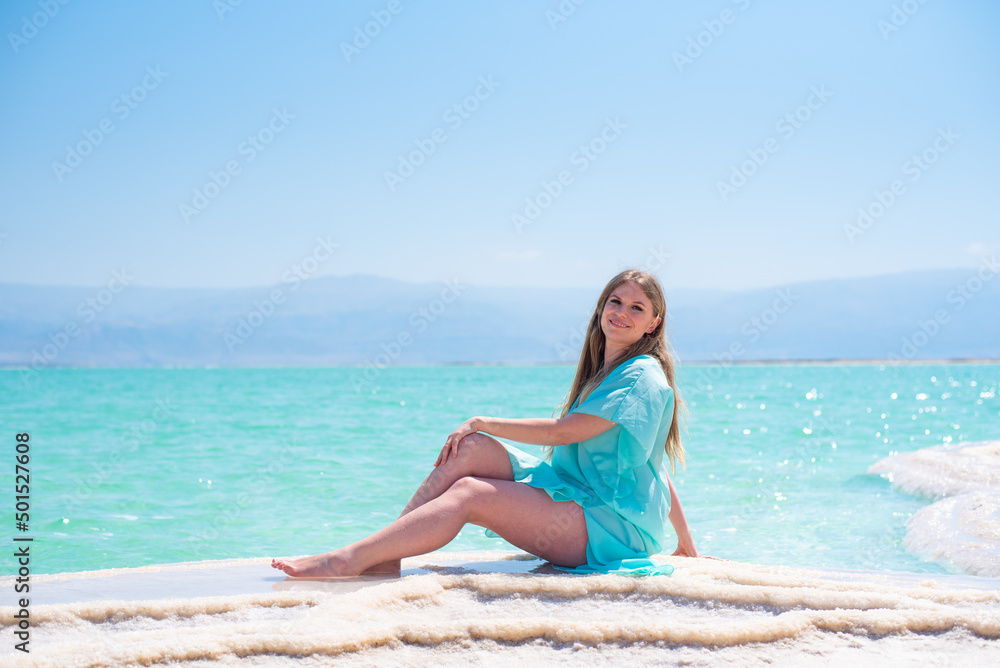 Slim woman in turquoise pelerine sits on salt island of Dead Sea on background sea and mountains on horizon. Treatment and recreation in Israel, salt and mud are good for health. Slim woman in turquoise pelerine sits on salt island of Dead Sea on background sea and mountains on horizon. Treatment and recreation in Israel, salt and mud are good for health.