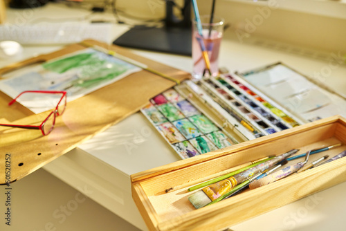 Watercolor drawing, glasses, watercolors in a box, brushes on the table, nobody