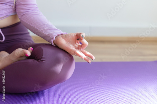 Young woman practicing yoga, relaxing in Lotus pose on mat. Padmasana exercise close up, mudra hand and feet close up. Girl wearing purple sportswear, meditating in yoga studio or at home