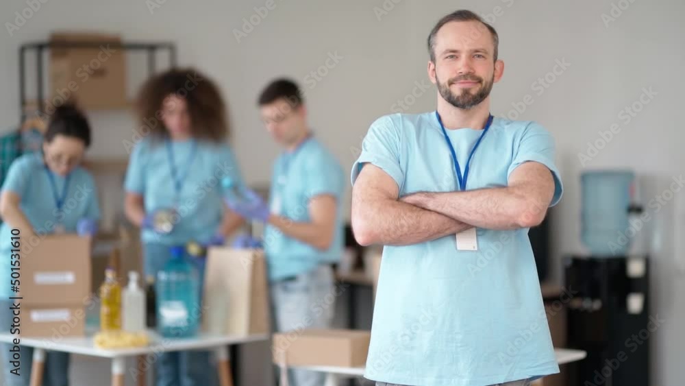 Smiling caucasian male volunteer in blue uniform and protective gloves standing with arms crossed. Team sorting, packing items in cardboard boxes Smiling caucasian male volunteer in blue uniform and protective gloves standing with arms crossed. Team sorting, packing items in cardboard boxes
