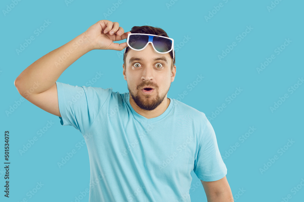 Young man surprised by something extraordinary. Happy handsome guy in blue T shirt lifts his sunglasses, looks at camera with funny shocked face expression and says WOW. Studio shot on blue background Young man surprised by something extraordinary. Happy handsome guy in blue T shirt lifts his sunglasses, looks at camera with funny shocked face expression and says WOW. Studio shot on blue background