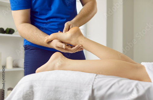 Male masseur therapist doing reflexology foot massage to young woman in modern beauty center. Cropped image of masseur therapist massaging legs of woman who is lying on massage table. Health concept.