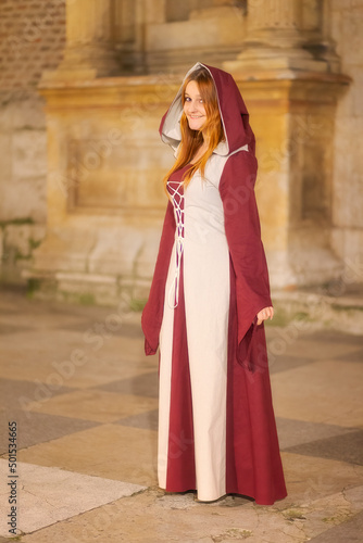 Smiling red-haired girl in long red and white medieval dress with a hood standing by a historic building at night