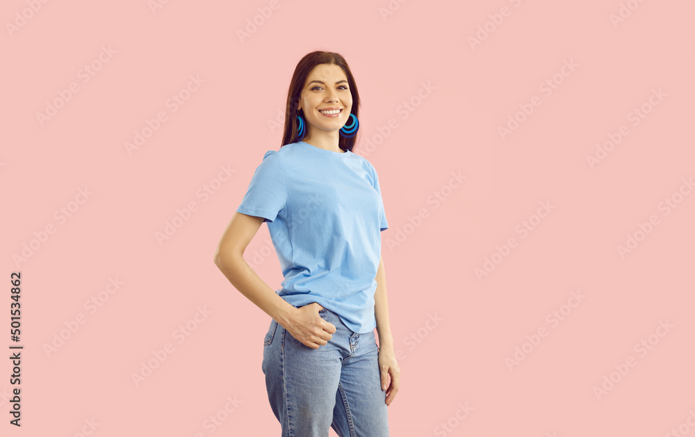 Studio shot of happy beautiful confident young woman wearing blue cotton T shirt, comfortable blue jeans, and pretty earrings standing isolated on light pink colour background. Casual fashion concept Studio shot of happy beautiful confident young woman wearing blue cotton T shirt, comfortable blue jeans, and pretty earrings standing isolated on light pink colour background. Casual fashion concept