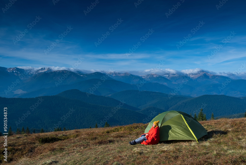A young girl sitting near the camp tent with mountains peaks and blue sky on background. Happy girl resting in campsite