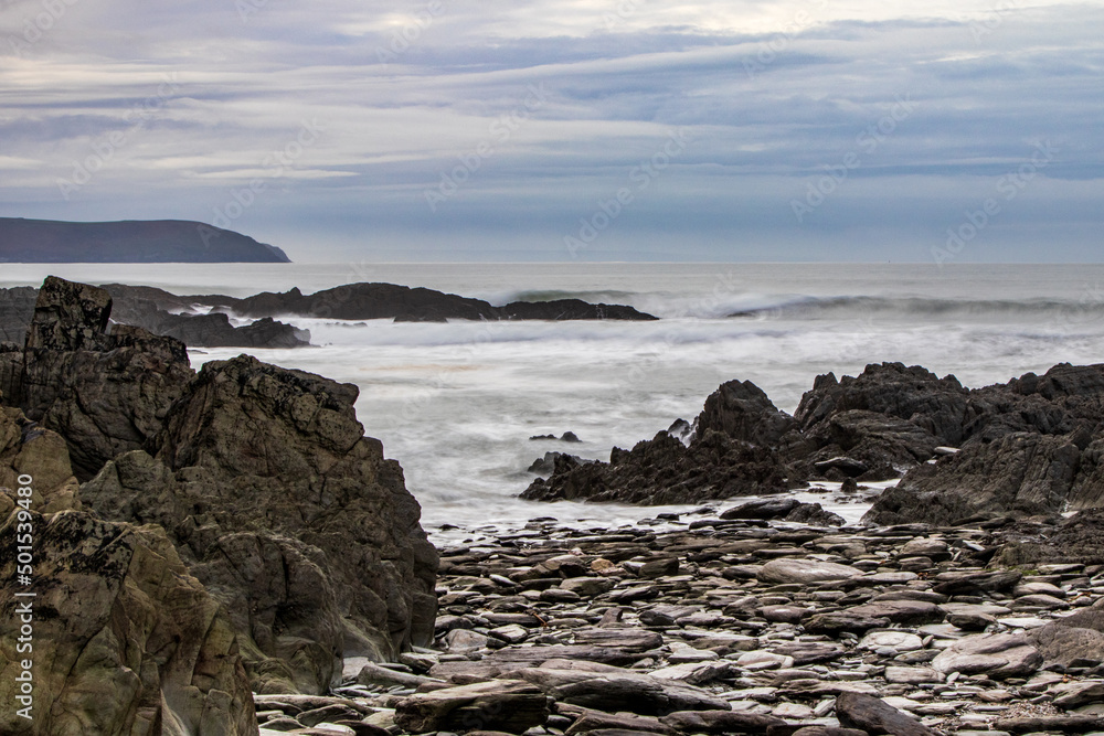 Fototapeta premium Stones and rocks on the beach, with the sea and a dark headland beyond.