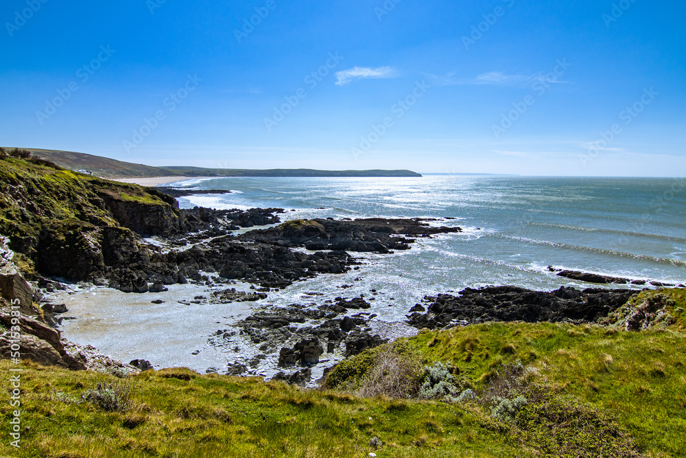 The view from Mortehoe towards Woolacombe Bay, on a glorious April day.  The sea is calm and sky is blue, with just a couple of fluffy clouds.  The green headland is visible in the foreground.