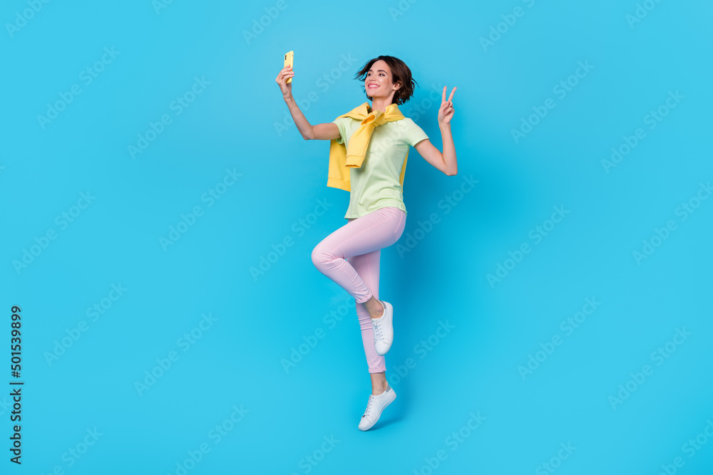 Full length photo of cute millennial brunette lady jump do selfie wear t-shirt pants shoes isolated on blue background