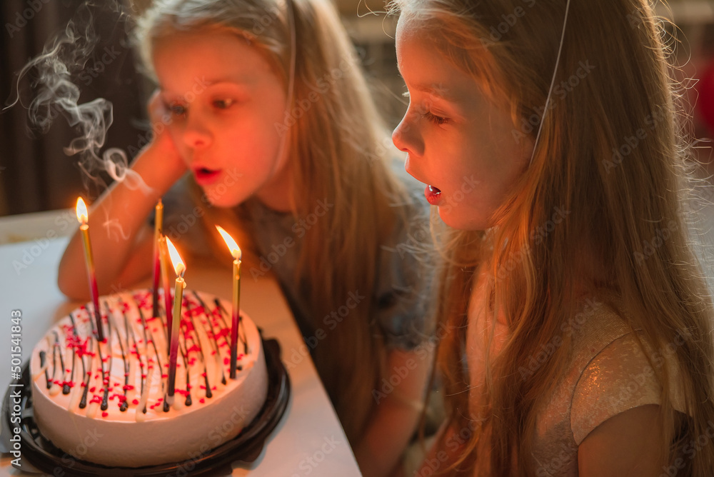 little girls, sisters, twins happily blew out the candles on the birthday cake at home festive cap. child birthday during illness, quarantine, isolation