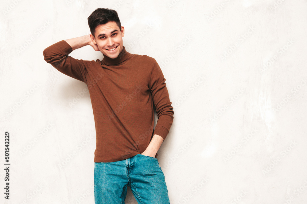 Portrait of handsome smiling model. Sexy stylish man dressed in brown turtleneck sweater and jeans. Fashion hipster male posing near grey wall in studio Portrait of handsome smiling model. Sexy stylish man dressed in brown turtleneck sweater and jeans. Fashion hipster male posing near grey wall in studio