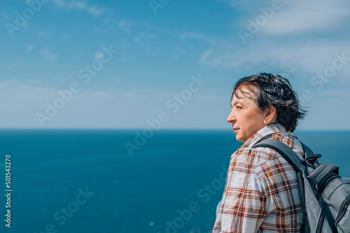 an adult woman with a backpack went hiking in the mountains near the sea in summer, a spring sunny day for health. pensioner, sat down to rest on a bench on top of a mountain, admires the sea