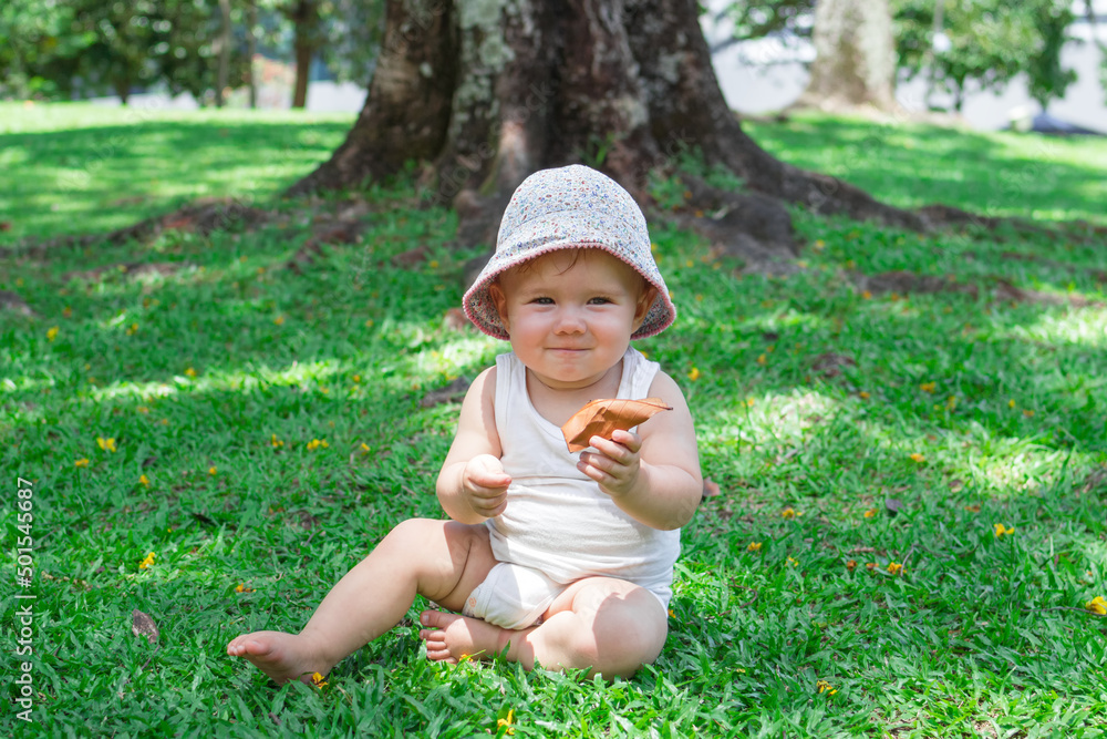 Adorable baby girl in white clothing and panama is playing with dry yellow leaves on grass. Summer sunny day in park. Adorable baby girl in white clothing and panama is playing with dry yellow leaves on grass. Summer sunny day in park.