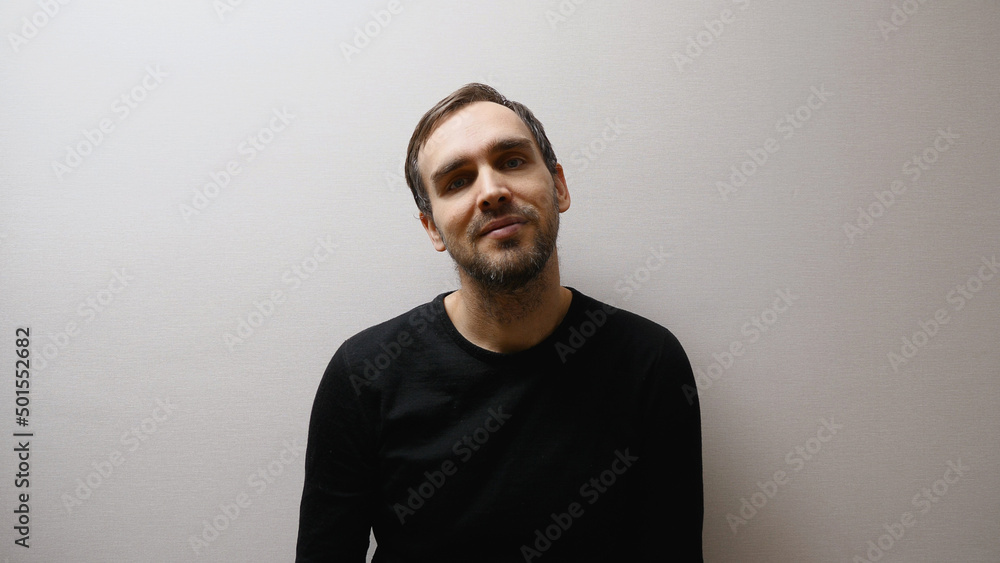 A handsome middle-aged Caucasian man in a black sweater, smiling as he stands in a room against a gray wall.