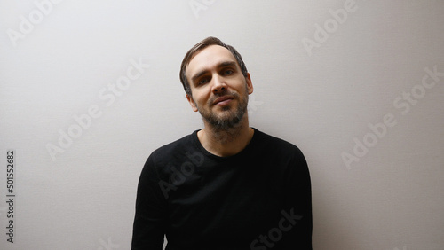 A handsome middle-aged Caucasian man in a black sweater, smiling as he stands in a room against a gray wall. © VeNN