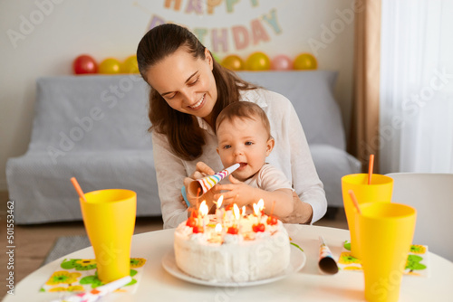 Portrait of mother with funny baby celebrating first birthday, cake is surprise for child, make a first wish, blowing party horn, posing at home with balloons and party inscription on background. photo