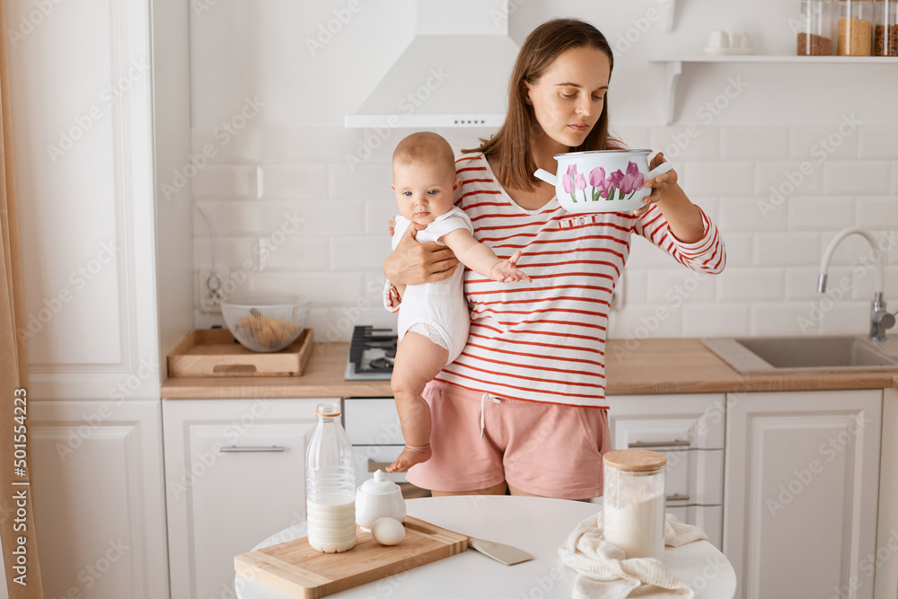 Portrait of young adult woman standing at the table in the kitchen with baby daughter in hands, being ready to cook or bake, holding pot and smelling what inside. Portrait of young adult woman standing at the table in the kitchen with baby daughter in hands, being ready to cook or bake, holding pot and smelling what inside.