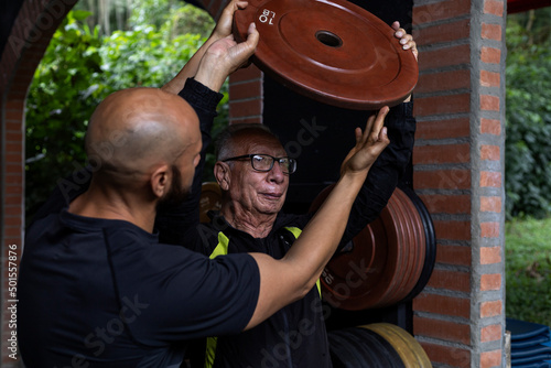 An older Latin American man and his trainer hold a weight disc in their hands that they use for rehabilitation and therapy exercises in the gym. Healthy lifestyle concept.