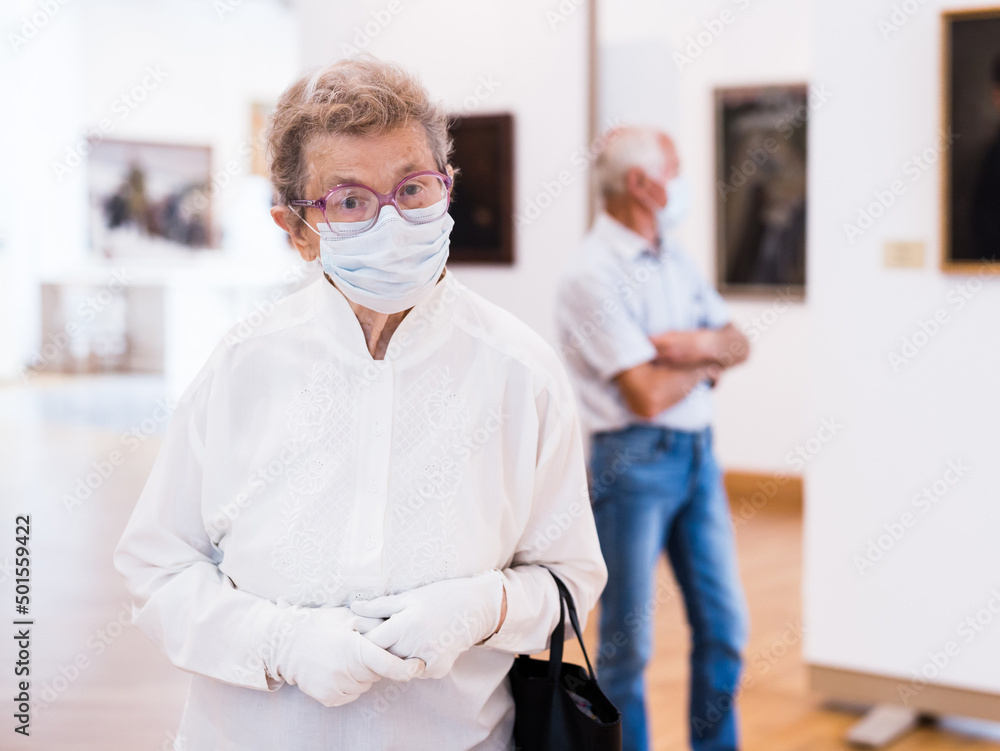 elderly European woman in mask protecting against covid examines paintings on display in hall of art museum elderly European woman in mask protecting against covid examines paintings on display in hall of art museum