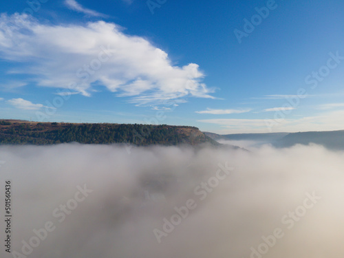 A thin morning fog covers the mountains. Green pines on the slopes of the mountains. Aerial drone view. 