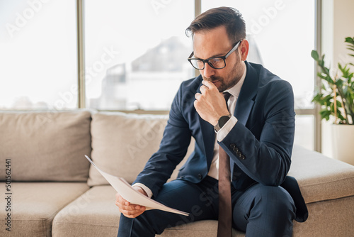 Foto Serious businessman reading documents while sitting on sofa against window