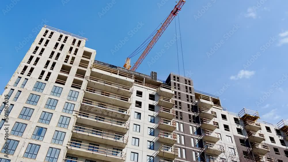Construction of the high floor house at sunny day, the worker in a protective helmet does a facade, construction cranes, the bottom view up, concrete blocks with windows Construction of the high floor house at sunny day, the worker in a protective helmet does a facade, construction cranes, the bottom view up, concrete blocks with windows