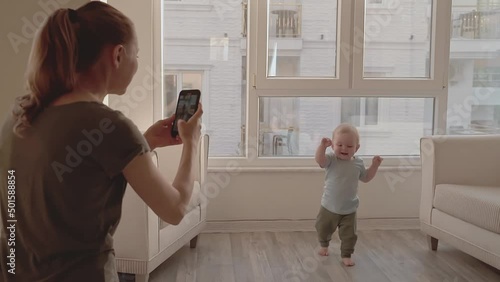 Young mother taking video of her adorable baby's first steps using smartphone, enjoying her motherhood. Mommy shooting video of her toddler son learning to walk for social media on cellphone camera