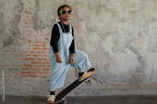 Little African kid boy with dreadlock hairstyle wear cool gold sunglasses, necklace chain, jeans bib and sneaker smiling and standing on skateboard on cement floor and brick wall on background photo