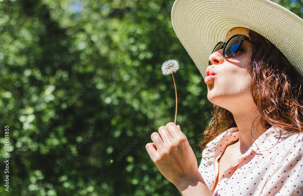 Young happy curly woman wearing hat and sunglasses blowing on a dandelion in nature. Young happy curly woman wearing hat and sunglasses blowing on a dandelion in nature.