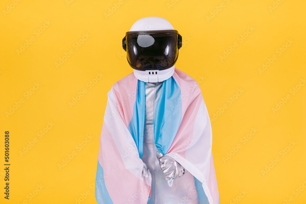 Gay man dressed as an astronaut with a helmet and silver suit, holding a flag of the trans community, on a yellow background. Gay, homosexual, trans, rights and gender pride concept. Gay man dressed as an astronaut with a helmet and silver suit, holding a flag of the trans community, on a yellow background. Gay, homosexual, trans, rights and gender pride concept.