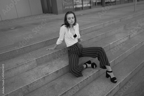 A young brunette girl in white shirt and striped trouser is sitting on the stairs
