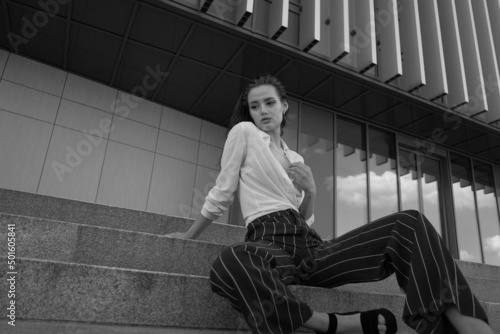 A young brunette girl in white shirt and striped trouser is sitting on the stairs